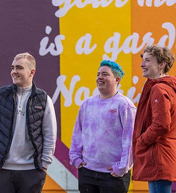 three young people standing smiling in front of a coloured mural